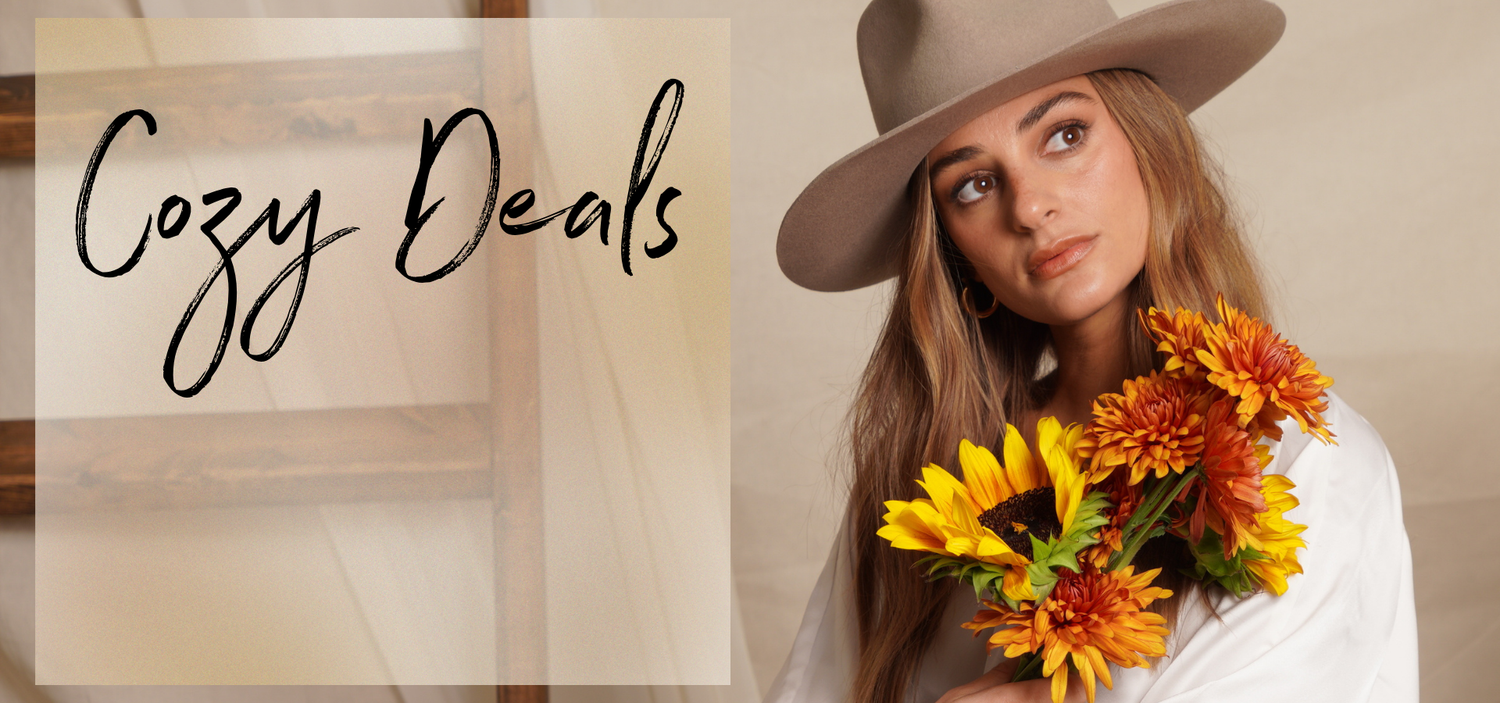 Our Boho model posing in a studio, wearing a white blouse, with her long dirty-blonde hair down down, as she's wearing a festival hat. She's holding a bunch of orange-yellow-red carnations with a single sunflower. A transparent square is superimposed on the left side of the screen with the words written on it in cursive (as if by fountain pen): Cozy Deals