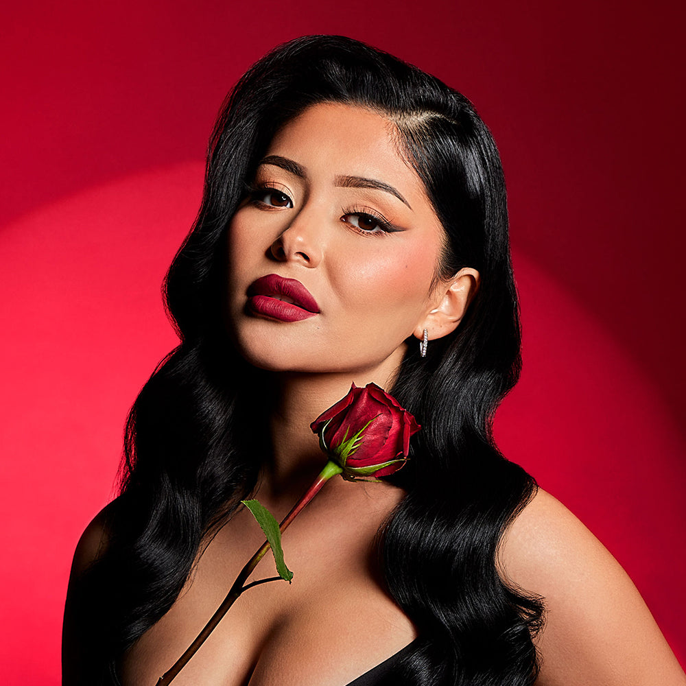 Vampy model with long black 1950's bombshell hair and ruby red lipstick. She's holding a rose beneath her face and is posing in front of a red backdrop with a spotlight pointed at it.