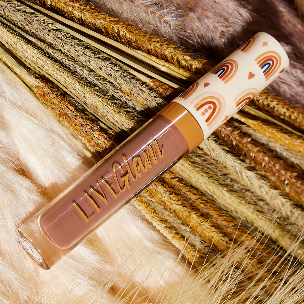 A bottle of the 'Dreamer' LiveGlam matte liquid lippie, with a cap featuring a boho-design with hearts, stars and skintone colored rainbows. The bottle rests on the autumn-themed bed of wheat and faux-fur