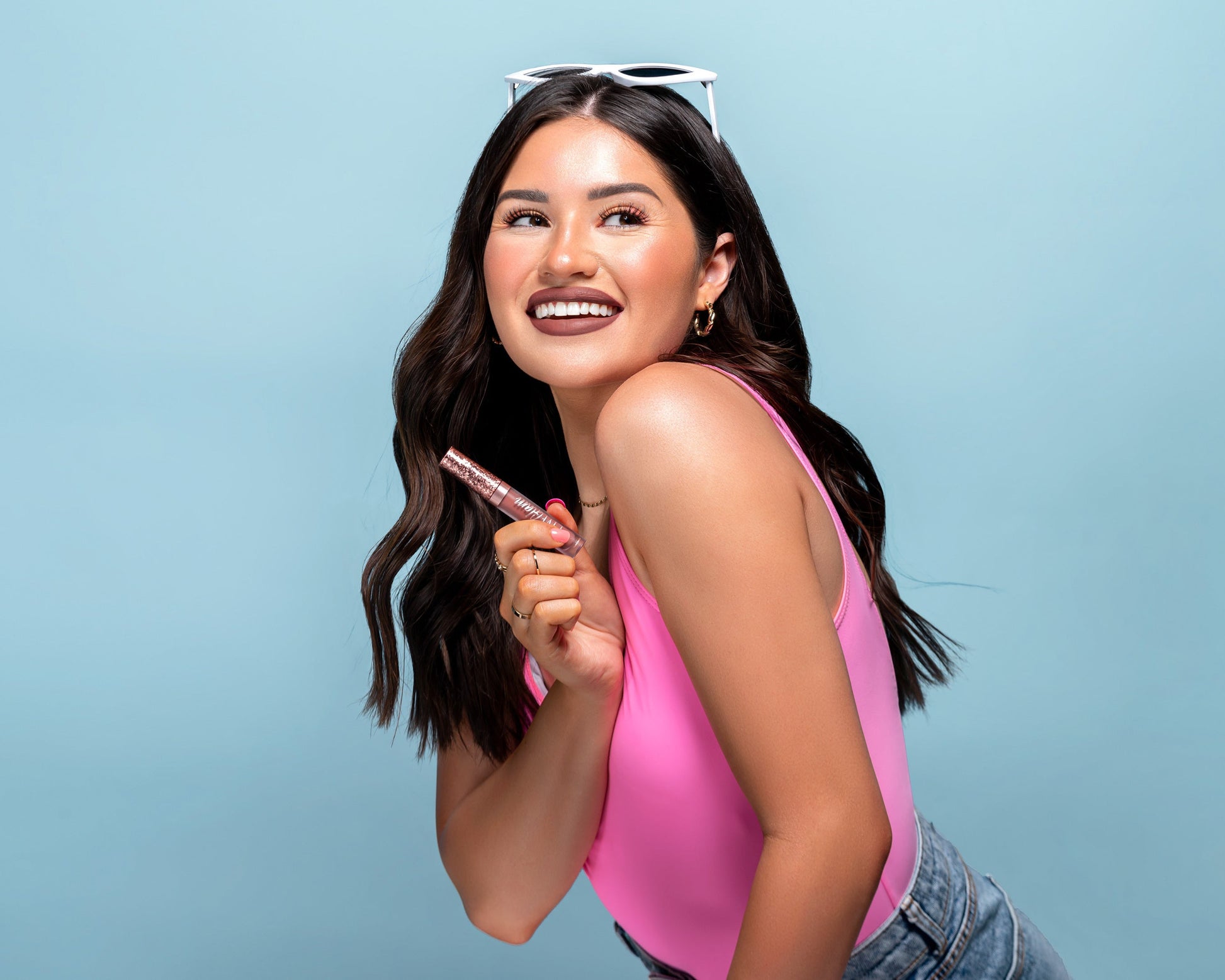 Woman in pink top and jeans and white sunglasses on her head, holding a HOTTIE matte liquid lippie in front of a light blue background