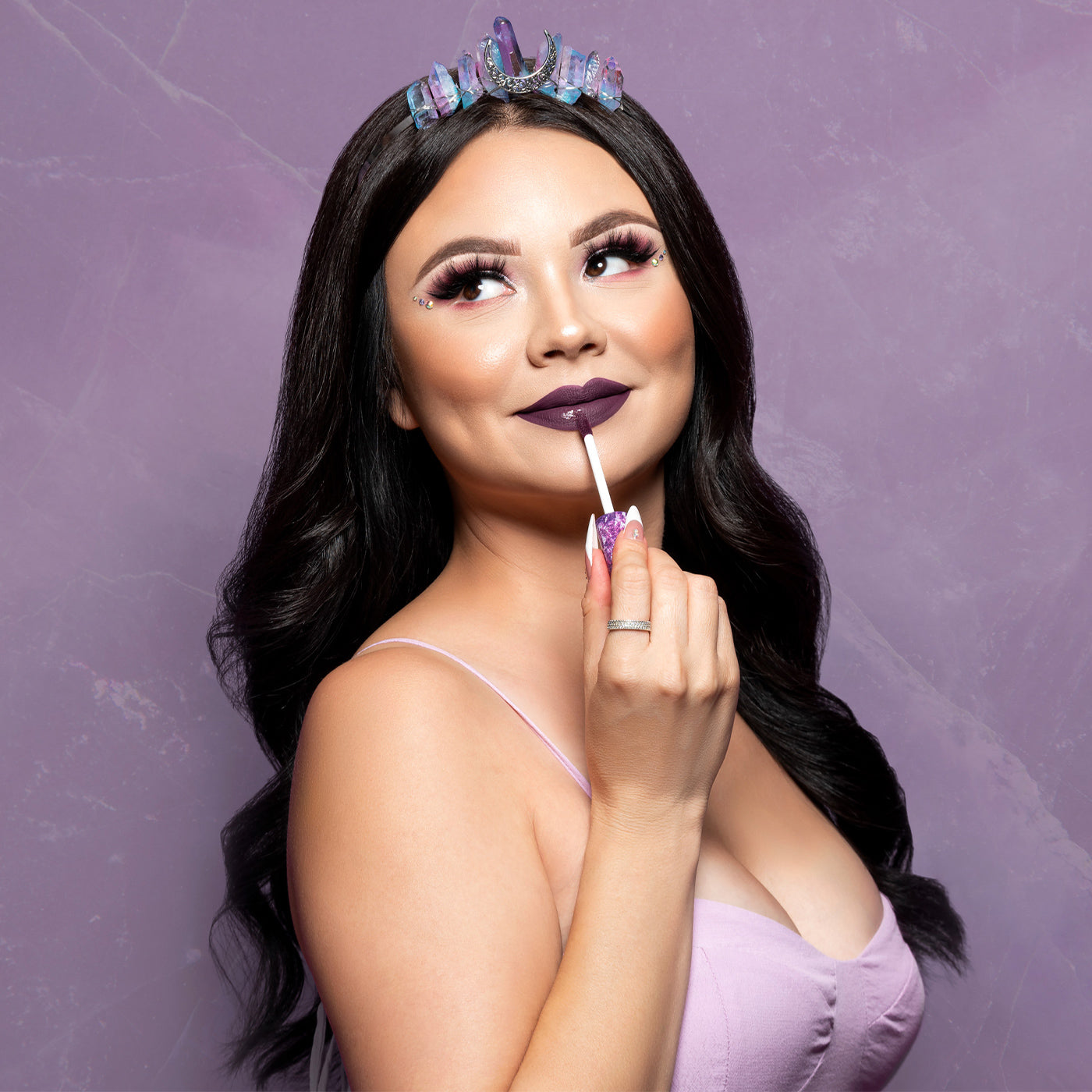 Standing in front of a light purple marbled background, a model is wearing a geode-and-gem tiara and applying the dark purple 'May the Quartz Be With You' liquid lippie with the applicator cap, as her eyes are looking up to her right (and showing off LiveGlam eyeshadow.