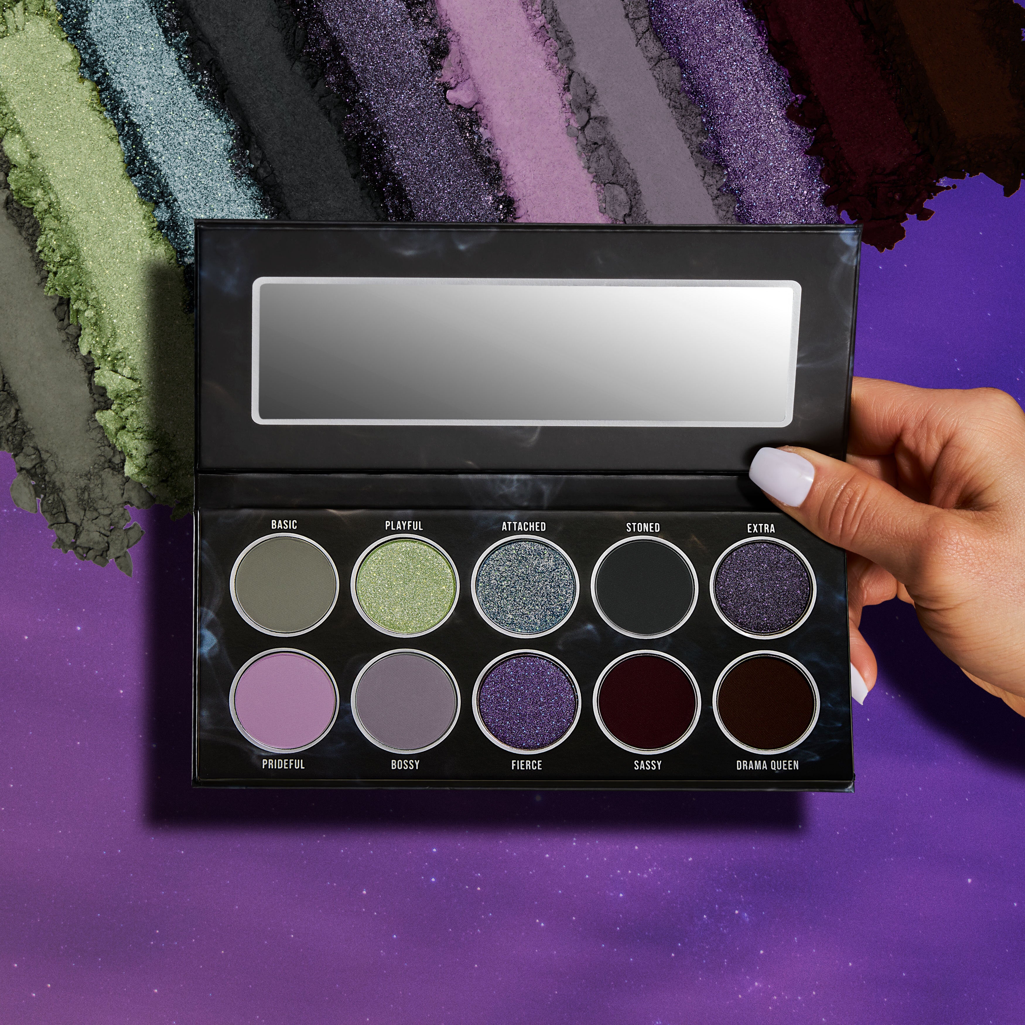 A hand with white nail polish holding a shadow palette in front of color swatches for each of the 10 shadow colors on a background of a purple sky with stars