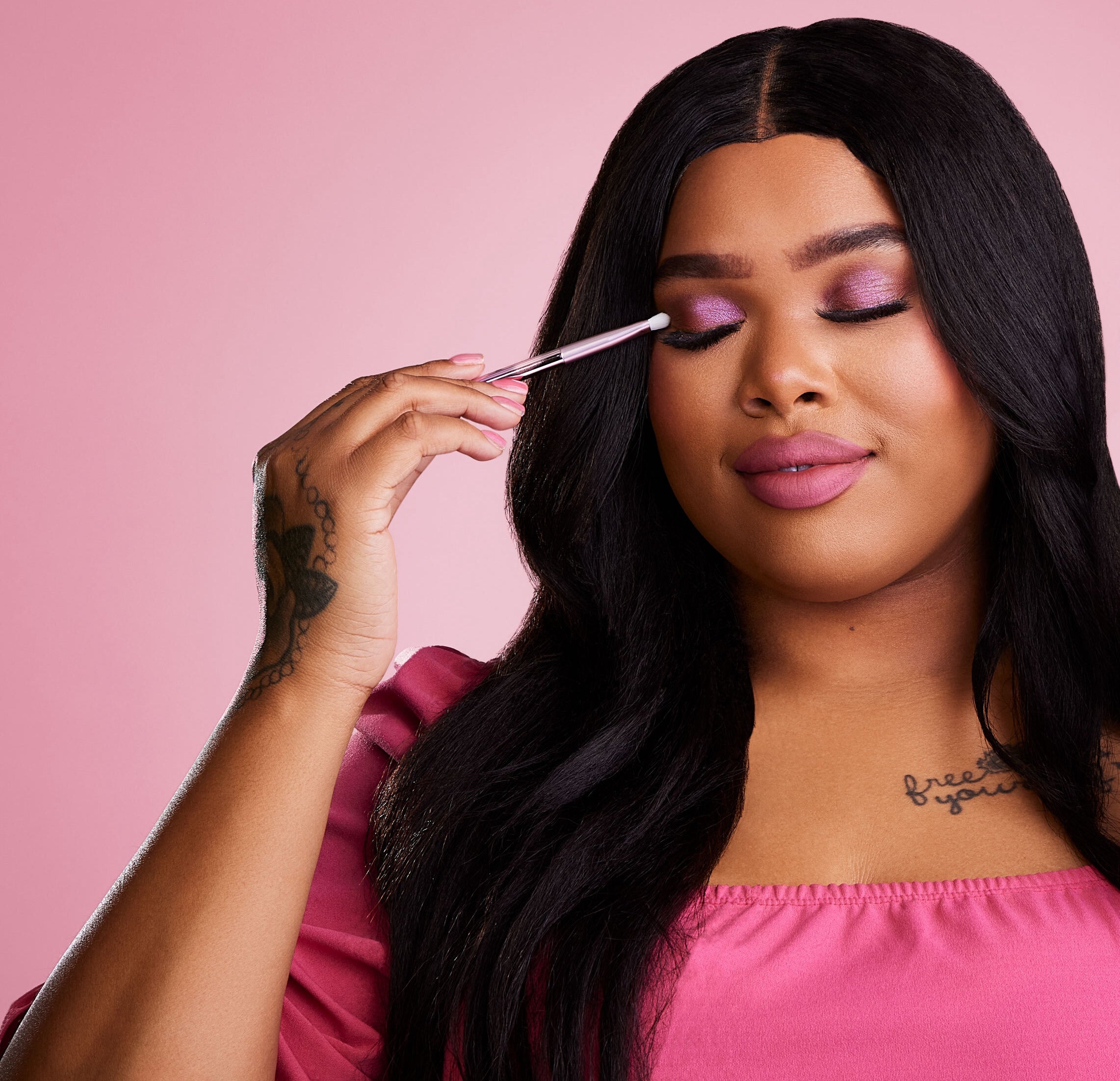 A model wearing pink and with pink nails holds a LiveGlam Brush #100 up to her closed eyelids, which have a sparkly pink shadow applied