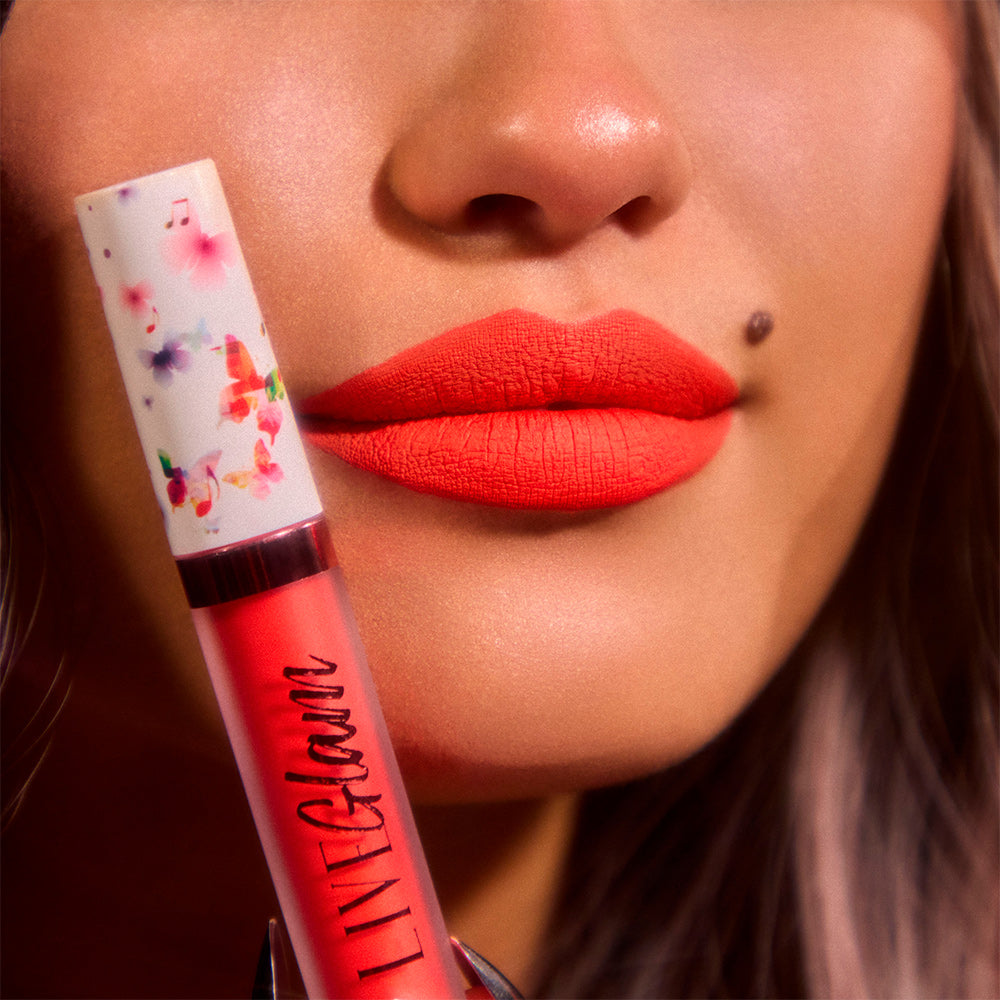 A close-up image of the lower face of our model, wearing LiveGlam red-orange matte liquid lippie 'Rose Over Bros' on her lips and holding a bottle of 'Rose Over Bros' in butterfly-music themed cap up beside her face.
