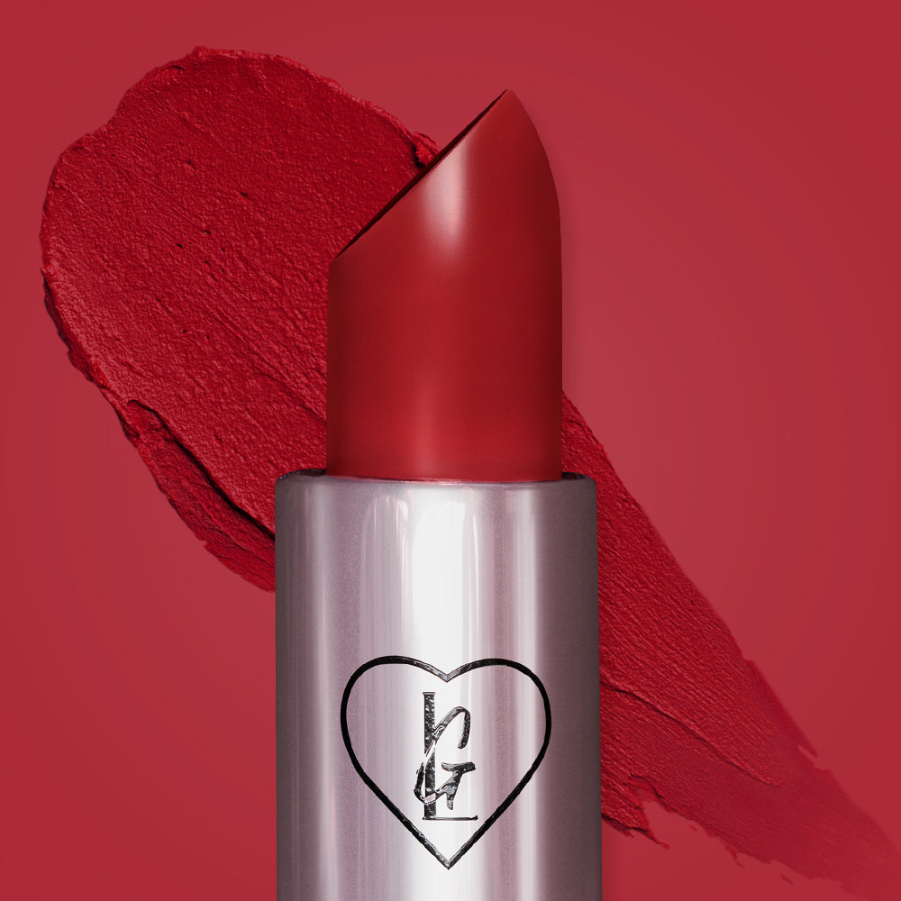 A close-up image of red Mami bullet lipstick in a true ruby red shade with a heart logo on the tube. Background shows a red backdrop as well as a thick red swatch from the bullet lippie
